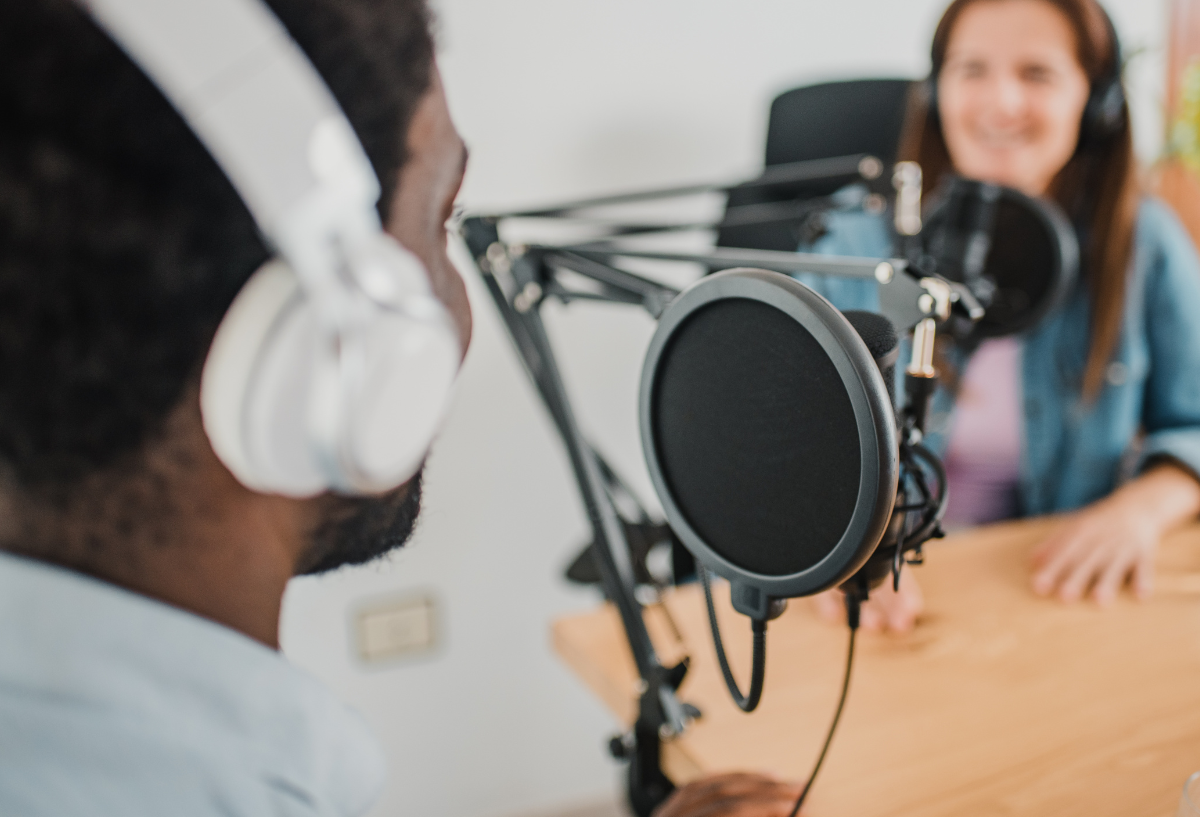 2 people speaking into microphones at a desk, wearing headphones