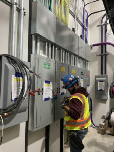 A field associate electrician works on breaker boxes at a construction site. The electrician is wearing a hard hat, safety goggles, gloves, and a high-visibility vest as part of their personal protective equipment (PPE). They are focused on wiring and making adjustments to the breaker boxes, surrounded by electrical tools and cables. The area is well-lit with overhead lighting, and electrical panels and exposed wiring are visible in the background. The electrician's attention to detail emphasizes the importance of safety and precision in electrical work on the site.