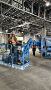 Inside a construction job site, workers are assembling a blue lift. The area is filled with construction materials and equipment. Several workers, wearing full personal protective equipment (PPE), are focused on the task. Each person is equipped with a safety harness, hard hats, gloves, and reflective vests. One worker is perched on the lift, while others are on the ground, handing materials or assisting with the assembly. The environment is well-lit with scaffolding and beams visible around the lift, showcasing a dynamic and active construction process.