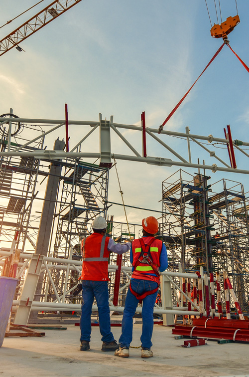 Two workers in hard hats and red shirts on a steel girder.