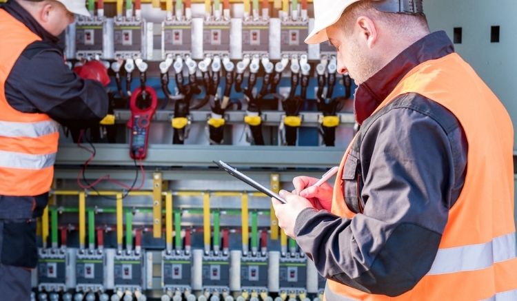 Workers in orange vests wiring a large electrical panel.